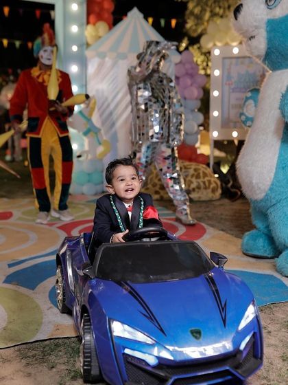 The birthday boy enjoying a ride in a mini car, with carnival entertainers like a juggler and a mirror man in the background.