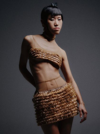 A still from the shoot, capturing the intricate texture of the gold outfit and the complementary choker and earrings. The lighting is dramatic, highlighting the metallic sheen.