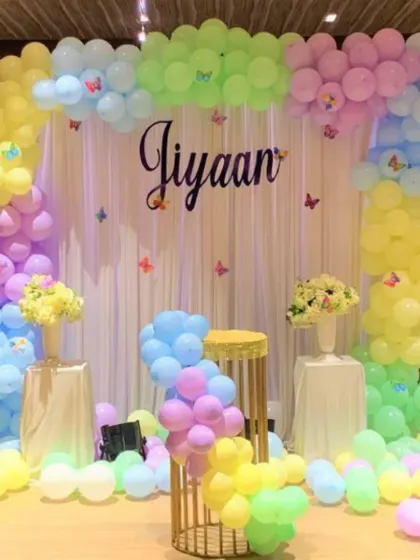 A soft and gentle pastel-themed decoration for a baby event. The backdrop is framed by a balloon arch in pastel rainbow colors with small butterfly accents.
