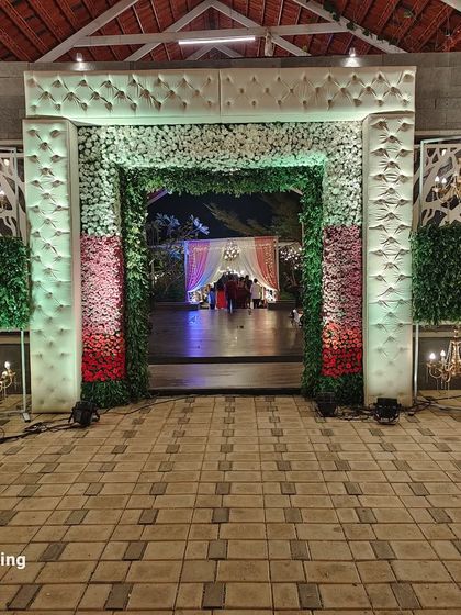A grand main entrance for a reception, featuring padded white frames, walls of green and red flowers, and elegant chandeliers.