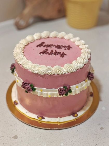 A lovely pink vintage-style cake with "Happy Birthday" written in a flowing script. The white buttercream frills and tiny piped roses give it a classic, celebratory feel.