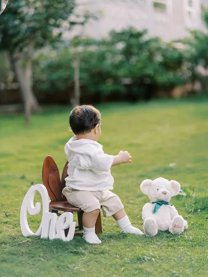 A sweet, candid shot from behind, showing the birthday boy with his "ONE" sign and teddy bear, looking out at the world.