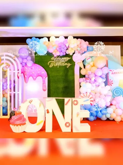 A sweet and colorful Candyland first birthday stage. The backdrop features a faux grass wall, pastel balloon garlands, and large props like a dripping ice cream cone and a donut, with "ONE" marquee letters in the center.