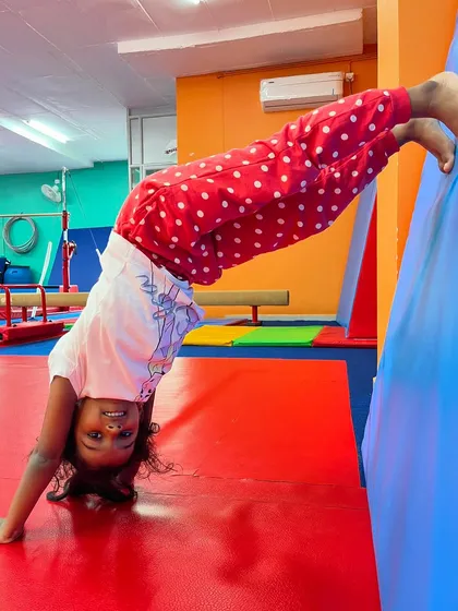 A girl practices a wall-assisted handstand, smiling at the camera. This is a great way to build confidence with inversions, using the wall for support to hold the position and build strength.