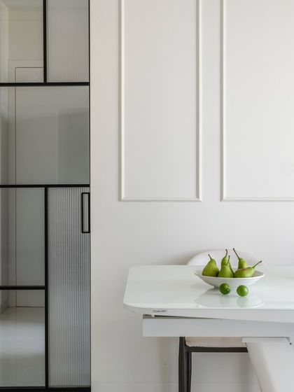 A detail shot from a minimalist interior project, focusing on the interplay of textures. The fluted glass of the door, the smooth white of the table, and the simple wall mouldings create a quiet, layered composition.