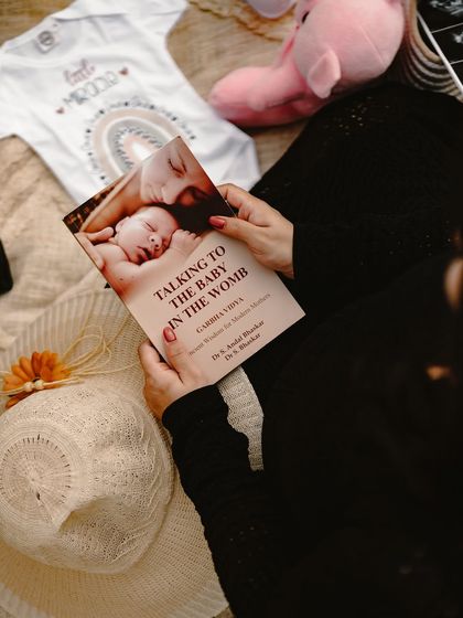 A cozy, detailed shot of the mom-to-be reading "Talking to the Baby in the Womb," surrounded by other props like a onesie and a hat. It shows the thoughtful preparation for motherhood.
