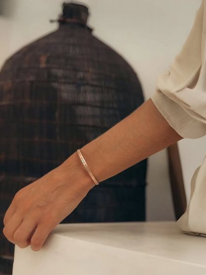 A woman wearing a rose gold cuff bracelet in a minimalist, artistic setting. The soft lighting highlights the warm tone of the rose gold finish.