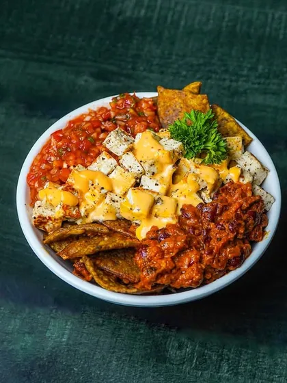 A bowl of loaded nachos with paneer, salsa, beans, and cheese sauce. The dark background and spotlight effect create a fun and indulgent vibe, perfect for social media.