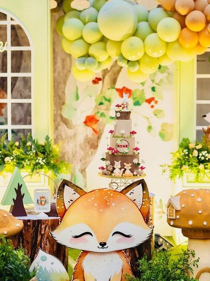 A detailed view of the woodland dessert table, with a cute fox prop in the foreground and the stunning tiered cake visible behind it. The scene is rich with texture, from the wooden stumps to the lush greenery.