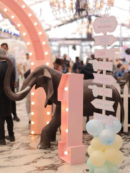 A charming corner at a pastel carnival party, featuring a light-up marquee number '1', a life-sized elephant prop, and a whimsical directional signpost pointing guests towards the cake, games, and fun.