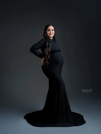 The classic black gown is a timeless choice for a maternity photoshoot. It creates a sleek, elegant silhouette that beautifully accentuates the baby bump against a simple grey studio background.