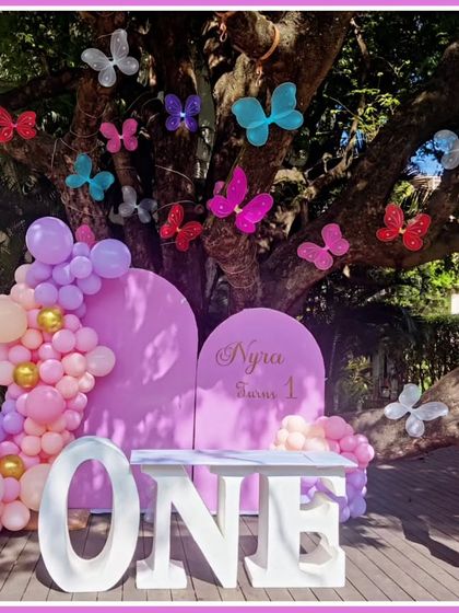 An outdoor butterfly-themed first birthday. The setup uses a large tree as a natural backdrop, decorated with colorful butterflies, and features a 'ONE' marquee table.