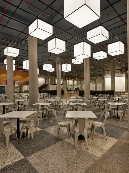 An industrial-style cafeteria with a checkerboard terrazzo floor, concrete pillars, and large, suspended box lights. The design is raw, spacious, and highly durable.