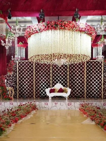 A truly grand reception stage, with a massive cylindrical canopy of hanging white and red flowers, set against a backdrop of red velvet and golden lattice screens.