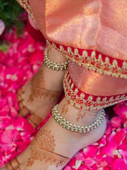 The rich red and gold embroidered border of the saree contrasts beautifully with the delicate henna on the bride's feet, set against a bed of rose petals.