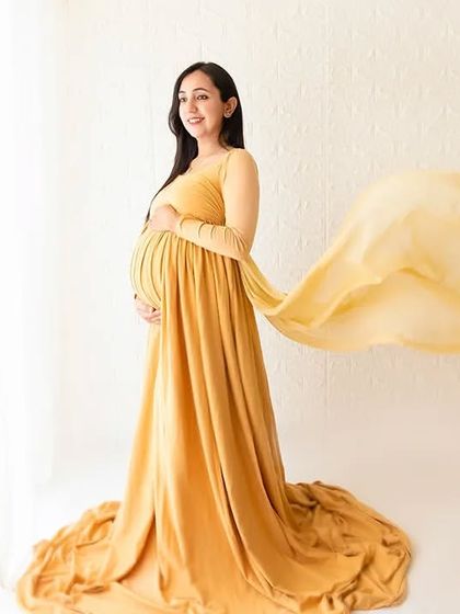 A side-profile maternity shot showcasing the baby bump in a beautiful yellow gown. The fabric flows behind her, creating a sense of movement against the minimalist white studio wall.