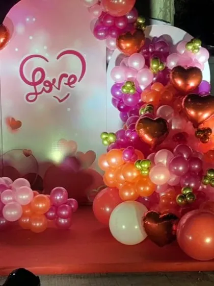 A close-up of our romantic Valentine's Day photo booth. The setup features a "Love" backdrop and a lush balloon garland in shades of red, pink, and orange, accented with heart-shaped foil balloons.