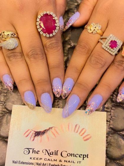 A beautiful set of long, almond-shaped nails in a soft lavender color. The tips are adorned with a mix of gold and iridescent glitter flakes for a magical, sparkling effect.