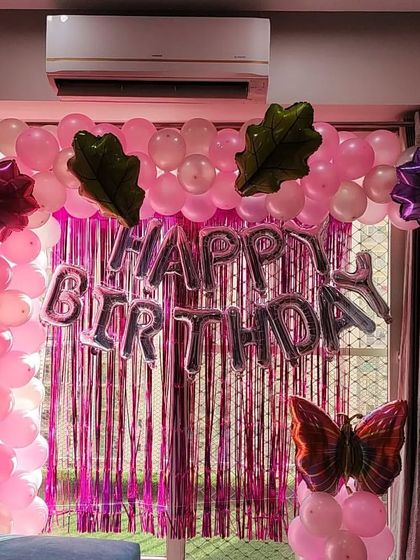 A pink-themed birthday setup in front of a window, using a foil curtain and various foil balloons.