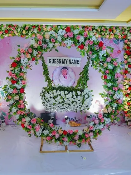 We also create beautiful traditional decorations for events like naming ceremonies. This stage setup features a lush floral arch in pink, white, and red, with a decorated cradle as the centerpiece for the baby.