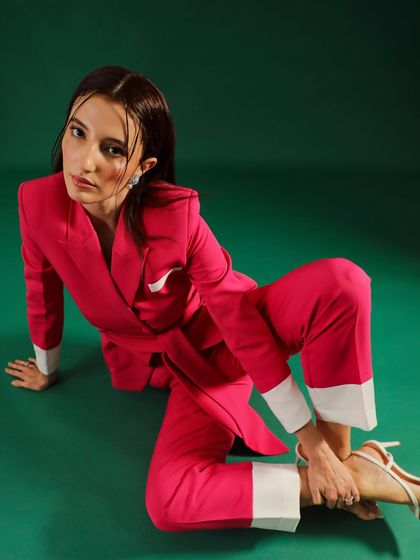A model in a bright pink pantsuit with contrasting white cuffs. The pose is unconventional and artistic, designed to make a statement in a fashion lookbook.
