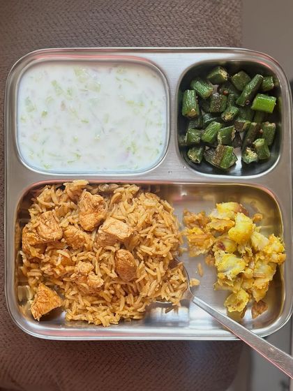 This plate features chicken biryani with sautéed okra, potato, and a side of cucumber raita.