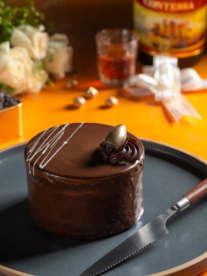 A rich chocolate truffle mini cake, styled with a liquor bottle and white roses to suggest a sophisticated flavor pairing.