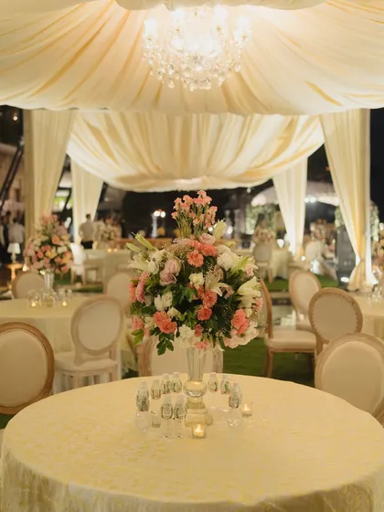 An elegant outdoor reception dinner setup under a draped canopy with a crystal chandelier. The tables are dressed in ivory linens with beautiful pink and white floral centerpieces.
