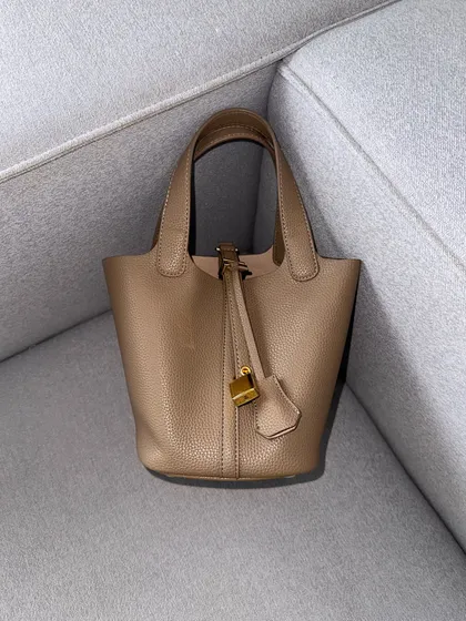 A clean shot of the HerCharm Bucket Bag in taupe, resting on a couch. This photo highlights the bag's soft structure and the signature lock and key charm.