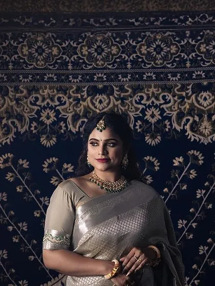 A wide shot of a model in a shimmering silver saree, posed against an ornate blue and gold carpet. The dramatic lighting and unique backdrop create a regal and theatrical portrait.