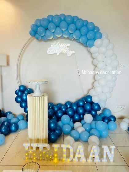A cool-toned birthday backdrop for a boy's party, using a circular frame with blue, chrome blue, and white balloons. The design is personalized with light-up marquee letters for the name 'HRIDAAN'.