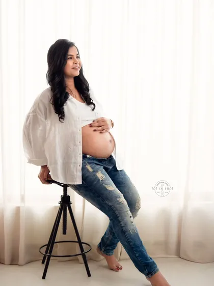 A relaxed and stylish portrait of a mom-to-be in jeans and a white shirt, seated against a brightly lit window for a clean, airy feel.