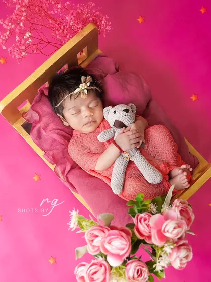 A newborn girl sleeps soundly on a miniature bed, hugging her teddy bear. The vibrant pink background and floral accents create a lovely, feminine portrait.