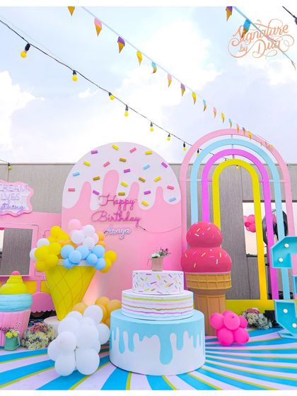 A full view of the ice cream-themed party setup. It includes a multi-tiered cake display, a giant ice cream cone, a sprinkle-covered backdrop, and a pastel rainbow arch, creating a complete visual feast.
