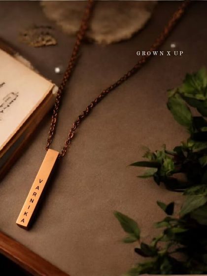 This flat lay shows a rose gold bar pendant with the name 'Varnika' engraved. The moody lighting and natural elements create an aesthetic that is both rustic and elegant.