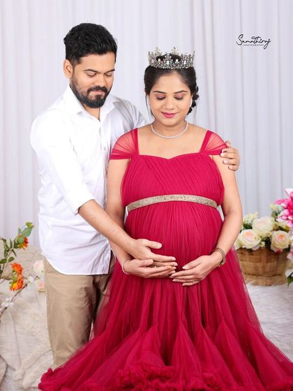 A sweet and tender moment between a couple. The mother-to-be, wearing a red gown and a crown, shares a loving glance with her partner in a beautifully decorated studio setting.
