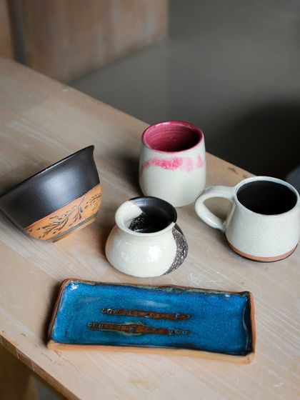 A stylish set of student work, featuring a rectangular tray with a brilliant blue glaze, a carved bowl, and mugs with contrasting colors.