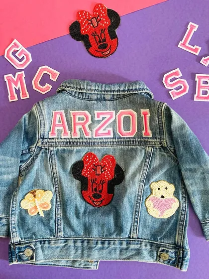 A flat lay of a custom denim jacket for 'Arzoi', showing the Minnie Mouse theme. The image also displays extra letter patches, illustrating the components we use for personalization.