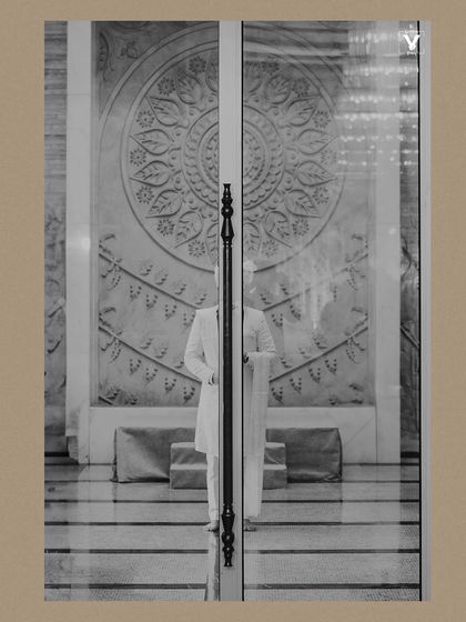 An artistic black and white shot of the groom seen through glass doors. The reflection and the architectural lines create a creative and visually interesting portrait.