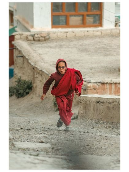 A young monk runs playfully down a village path, his red robes a splash of color against the earthy tones of the buildings.