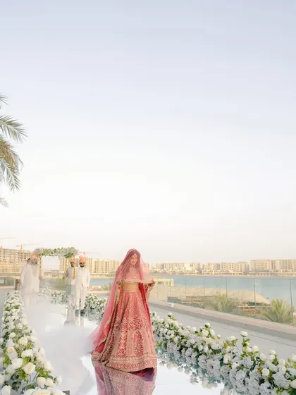 The bride's solitary walk to her forever. This wide shot captures the grandeur of the moment, her long pink veil trailing behind her on the mirrored aisle, with the Abu Dhabi skyline as a witness.