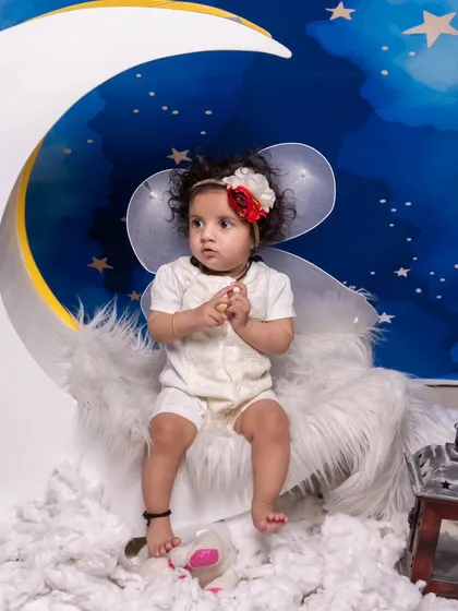 A dreamy 'on the moon' setup with a starry night backdrop and fluffy clouds. The baby is dressed as a little angel, sitting on a crescent moon prop.