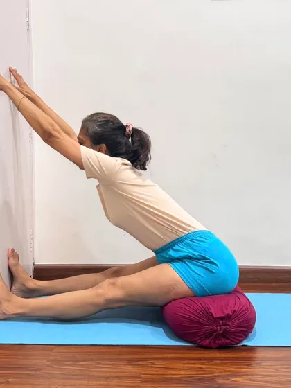 Seated on a bolster with feet against the wall, I demonstrate an active stretch in Dandasana. The bolster elevates the hips to maintain a straight spine, while the wall provides resistance to lengthen the hamstrings. This is a foundational pose for preparing the body for deeper forward bends.