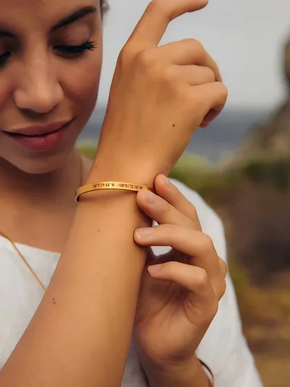 A woman adjusts a delicate gold cuff on her wrist, engraved with the coordinates of a special place. It's a subtle, meaningful way to carry a cherished memory with you always.