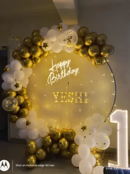 An elegant gold and white ring backdrop for a first birthday. The setup includes a neon 'Happy Birthday' sign, personalized name letters, and a light-up number '1'.