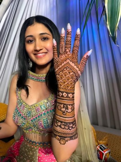 A smiling bride proudly displays her full-hand mehandi. The design extends up the arm with classic checks and floral patterns, ensuring she looks perfect from every angle.