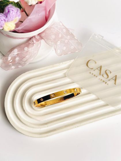 A single gold bangle placed on a textured tray. This minimalist approach draws all attention to the product, highlighting its classic design and finish.