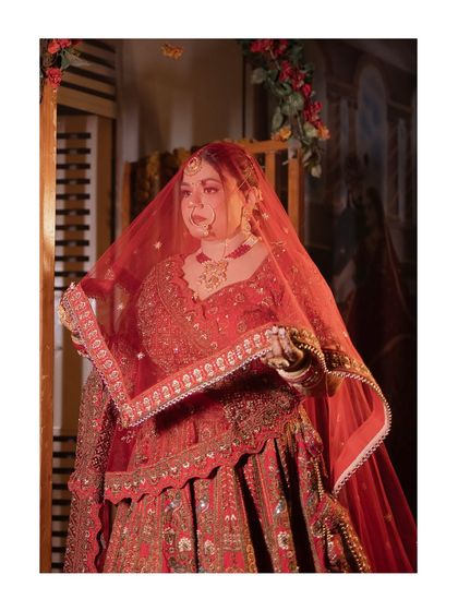 A delicate and soft portrait of the bride adjusting her red veil. The focus is on her gentle expression and the beautiful details of her nath and lehenga.