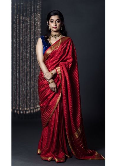 A full-length studio portrait of a model in a vibrant red saree with a contrasting blue blouse. The dark background and elegant pose highlight the richness of the festive attire.