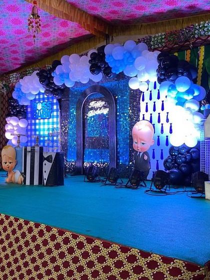 A Boss Baby first birthday stage under a tent, featuring a sequin backdrop, cloud-like balloon clusters, and character cutouts, including one peeking out of a tuxedo-themed box.